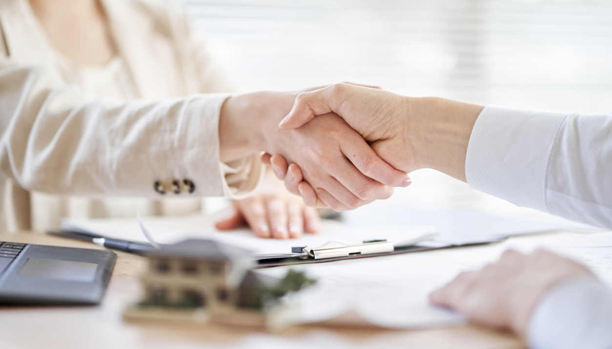 The image depicts two individuals shaking hands over a desk, signifying an agreement or a deal being finalized. On the desk, there are various items including a laptop, a clipboard with documents. The atmosphere is professional, and both individuals are dressed in business attire.
